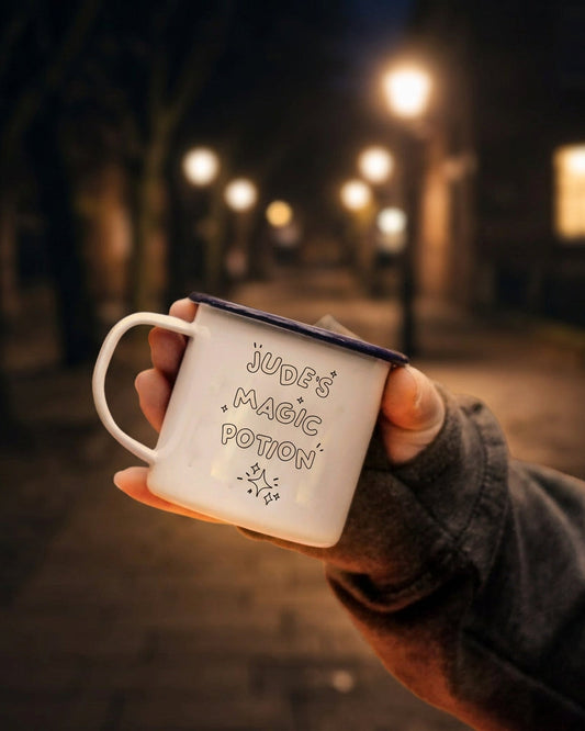 A hand wearing a dark gray glove holds a white enamel mug with a dark blue rim. The mug features the text 'JUDE'S MAGIC POTION' in a handwritten-style font with small star illustrations.